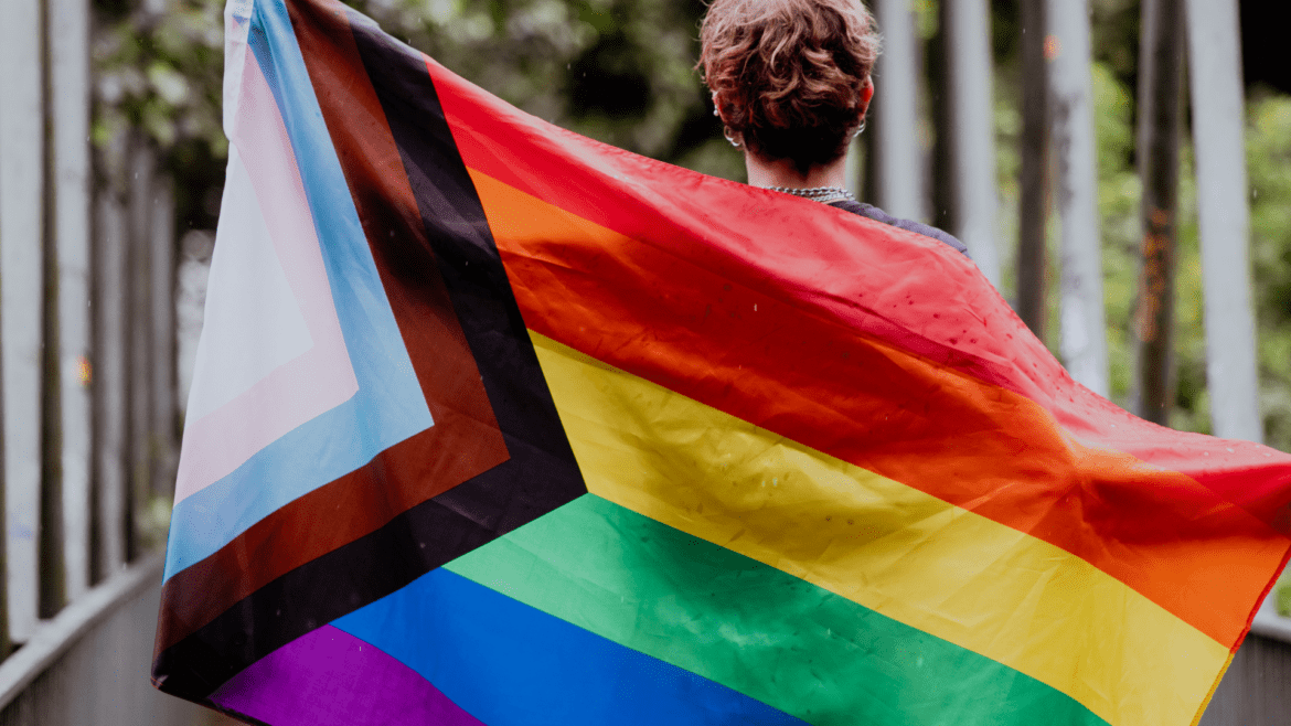 Lyhythiuksinen henkilö pitelemässä Pride-lippua selkänsä takana. Lipussa on muutamia sadepisaroita.
