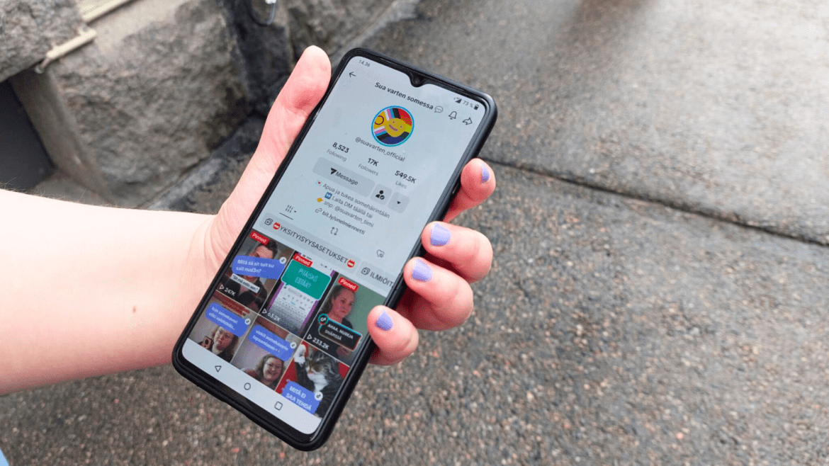 Käsi pitelemässä älypuhelinta, jossa on auki Sua varten somessa Tiktok-profiili. Taustalla asflattinen maa ja kivinen seinä.